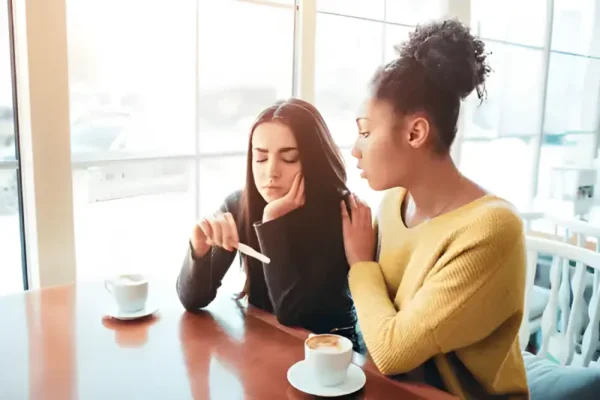 friend consoles a pregnant friend while looking at a pregnancy test in a coffee shop